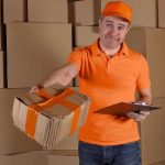 Man in orange uniform delivering heavily damaged parcel to customer. Brown cartons background. Unprofessional work and regret concept